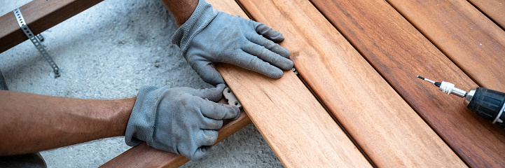 Male hands in protective working gloves placing a plastic spacer bewteen wooden planks as he lays new outdoor patio. Wide view image.