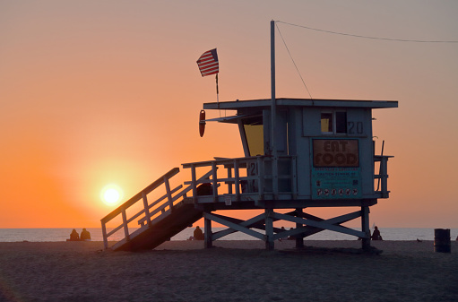 View of a lifeguard station on the Santa Monica Beach in Santa Monica, California right before sunset. Visible are the building itself, an American flag attached to the top, the setting sun, the Pacific Ocean, and several beachgoers sitting on the sand.