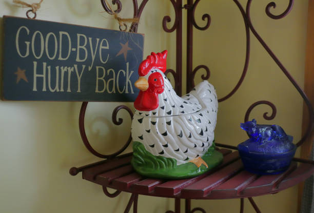 A charming vintage rooster-shaped ceramic cookie jar rests on a red bench next to blue glass salt cellar shaped like a wildcat. On the wall nearby hangs a wooden dark blue sign that reads “Good-Bye -- Hurry Back.” Yellow walls, afternoon sunlight.