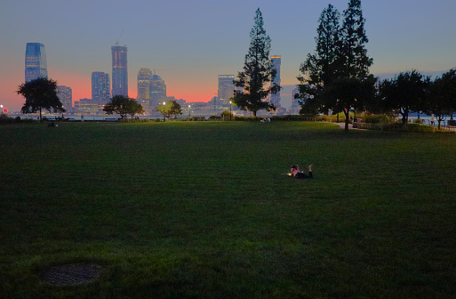 View of a Manhattan park near the Hudson River in New York, New York. Camera is facing west at dusk. In foreground a girl lies on the grass studying her smart phone, screen lighting her face.
