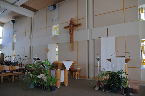 View of the altar of a modern American Catholic Church in Los Angeles. Visible are a crucifix hanging on the wall behind the altar, an ambo (podium) with microphones at right, a celebratory table left of center, an area for musicians in the left toward the back, and seats at the left. The scene is bathed in afternoon sunlight streaming in through windows.