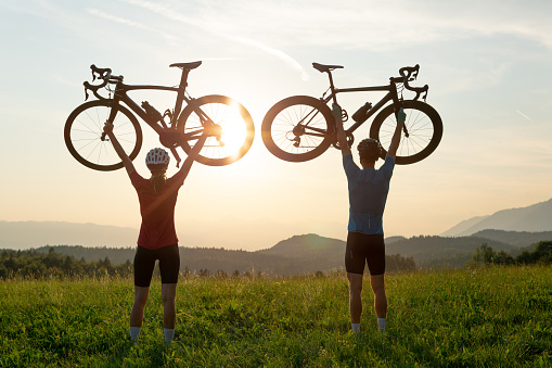 Two cycling racer silhouettes reach the goal and lift bikes above their heads and celebrate, with a mountain sunset view.