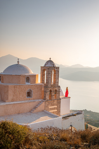 Young woman in a red dress watching the sunrise on the roof next to the bell tower and dome of Traditional Greek Orthodox church in Plaka Village on Milos Island, Greece