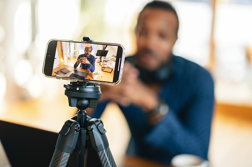 Portrait of an adult content creator speaking to a smartphone camera on a tripod, recording a video in a cozy home setting.
