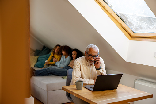 Senior adult man having a phone conversation and using laptop at home office table, with women and children relaxing in background.