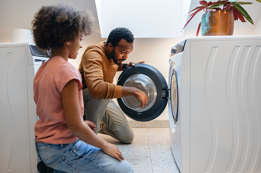 A caring African-American father shows his young daughter how to use the washing machine in a domestic setting.