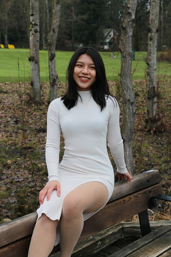 A Taiwanese woman sitting on the top of a wooden bench in a public park.  She is wearing medium length, black, straight hair, makeup, a big smile and a white long sleeved dress.