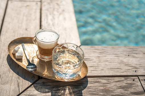 A serene setting displaying a glass of water and a cup of coffee on a golden tray, resting on a wooden deck by the crystal clear waters of a pool, suggesting a leisurely vacation moment at a villa.