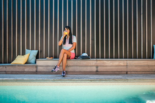 A mature Hispanic female relaxes by a pool, taking a leisurely break with a sandwich on a villa's comfortable outdoor bench, embodying the serenity of vacations at a luxurious rental retreat.