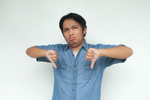 A young Asian man in a blue shirt  looking unhappy and angry showing rejection and negative with thumbs down gesture. Bad expression. Isolated in gray background.