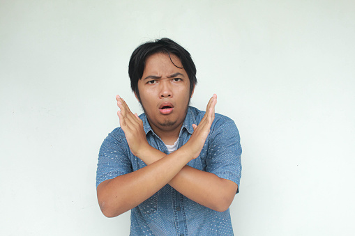A young Asian man in a blue shirt  looking unhappy and angry showing rejection and negative with crossed arms gesture. Bad expression, say no. Isolated in gray background.