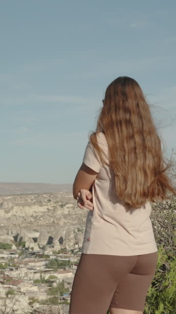Vertical video. A young woman stands at the top and admires the natural ancient park of Goreme from a high mountain, the camera zooms in on her.