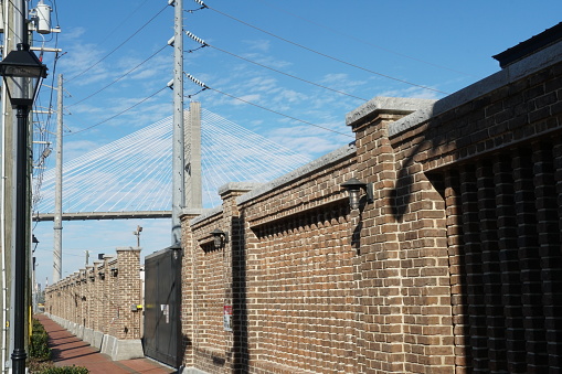 Savannah, GA, USA 01 01 2024: Sidewalk with old wall built of brown bricks in southern style with historical street lamps and electric pillars around in the American city of Savannah. Behind is Talmadge Memorial Bridge.