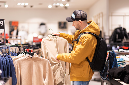 Bearded Caucasian young man wearing backpack chooses clothes in store. Concept of shopping and discounts.