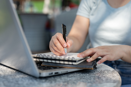 A close-up shot of a woman writing something in her notebook at an outdoor table, working remotely. taking notes, making list, listing ideas, jotting