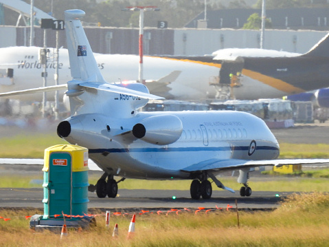 A Royal Australian Air Force Dassault Falcon 7X plane, registration A56-002, used for VIP transport of government ministers and others, is taxiing to the parking area of Sydney Kingsford-Smith Airport after arriving from Canberra.  In the foreground is a portable toilet.  In the background is a UPS Boeing B747-44AF plane parked at the international freight terminal. This image was taken from Shep's Mound, Ross Smith Avenue, Mascot, on a hot and sunny afternoon on 3 February 2024.