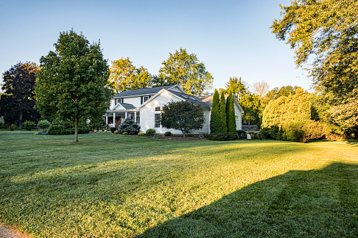 Early morning sun just after dawn under a completely cloudless pale blue sky. In the middle of September, with all of the landscaped plants and trees still fully dressed in lush, late summer foliage, it's peaceful and quiet this morning around this suburban Rochester, New York State colonial style house.