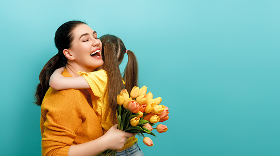 Happy women's day. Child is congratulating mom and giving her yellow flowers. Mum and girl smiling on teal background. Family holiday and togetherness.