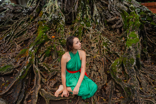 Serene woman in green dress sitting on the roots of tropical tree
