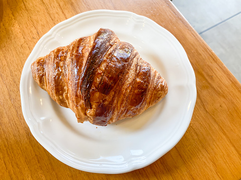 A plain butter croissant on white ceramic plate on wooden table. Concept for breakfast, restaurant and eatery.