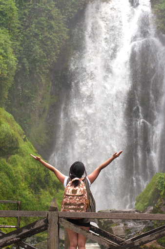 A lone traveler enjoys a moment of tranquility as she stands with arms outstretched facing a powerful waterfall surrounded by vibrant green foliage. High quality photo