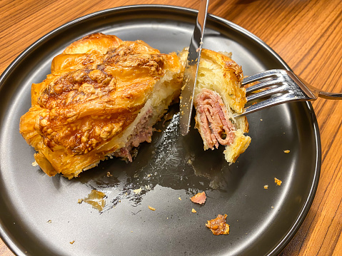 Someone was slicing a croissant filled with meat and cheese using a knife and fork on black ceramic plate. Concept for breakfast, restaurant and eatery.