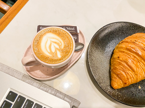 Hot Cappuccino Coffee with latte art in a glass next to croissant bread and laptop keyboard on white table. Caffe Latte or Coffee Milk Drinks and bread. Concept for relaxing time, working and business.