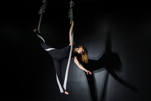 Play of light and shadow creates feeling of a theatrical performance. Sports activity. Fly yoga. Girl in a white hammock on a black background shows aerial acrobatics. Gymnastics, circus, under dome.