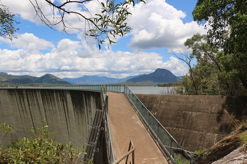 Moogerah Dam's solid concrete double curvature arch wall. Path across top of dam wall, tourists in the distance. Mountain range beyond.