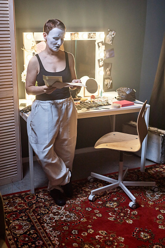 Vertical full length portrait of young woman as mime artist rehearsing backstage in theater standing by makeup table and preparing for performance