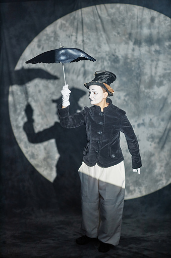 Vertical full length portrait of classic mime performer acting on stage in spotlight and holding umbrella