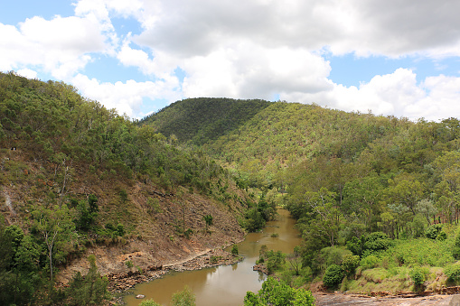 Downstream from Moogerah Dam, Reynolds Creek meanders through rocky, tree-lined banks. Muddy from recent controlled release. Semi-overcast day, shadows of clouds dapple the hillsides surrounding.