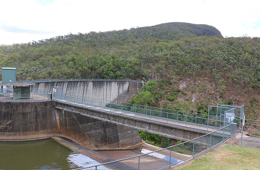 Regional 'Double Curvature Arch Dam', Moogerah Dam. Shown near capacity, almost at spillover level. A few tourists walk along the wall in the distance. Rugged hills with gumtrees surround.