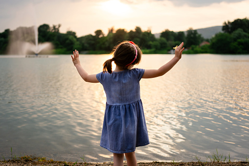 Rear view of a little girl with outstretched arms stands by a lake on a summer day.