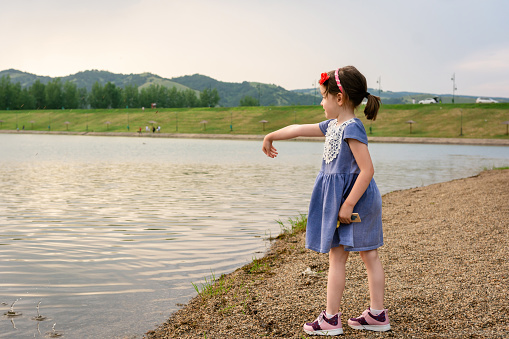 Side view of a little girl throws pebbles into a lake on a summer day.
