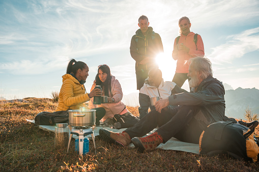 A diverse group of friends takes a break from hiking, sipping tea together at the scenic hilltop.