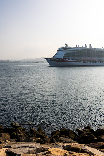 La Coruna, Spain 05 October 2023: The P and O Cruises' IONA, a modern leviathan of the seas, is pictured anchored in the calm waters. The foreground shows rugged rocks at the water's edge, providing a natural contrast to the ship's engineered grandeur. The gentle morning light bathes the scene, lending a serene glow to the ocean and the vessel. This peaceful maritime setting underscores the leisurely pace of cruise travel.