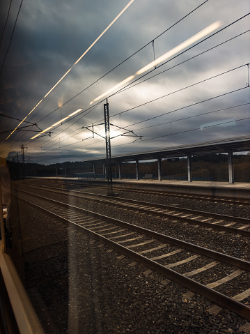 The convergence of the real and the reflected creates a captivating scene from the perspective of a train passenger. Overhead, the fluorescent lights inside the carriage streak across the window, mirroring the parallel rails below that stretch into the distance. The overcast sky, a canvas of shifting grays, imbues the moment with a sense of anticipation and travel's quiet introspection. This intersection of interior and exterior worlds encapsulates the transient nature of a journey just begun.