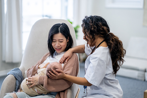A new Mother attends a Lactation Consultant appointment as she looks for guidance with breastfeeding her daughter.  The practitioner is dressed professionally in scrubs as she calmly helps with positioning and latch.