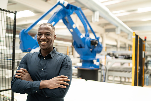 A smiling African-American man is standing with his arms crossed and smiling at the camera, in the background a factory can be seen with a large automatic robot arm