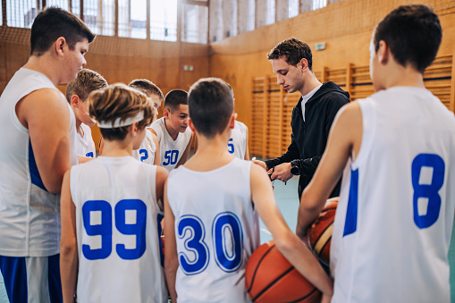 A young basketball coach is standing with his junior team and discussing tactics and strategy on court.Basketball kids standing on court with their trainer and working on game plan on basketball court