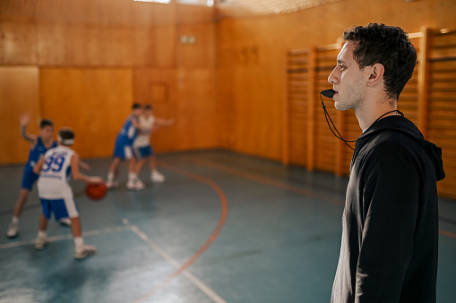 Profile view of a basketball coach on basketball court using a whistle during the game practice. A trainer is using whistle to stop a game practice because of foul. A coach using whistle on court.