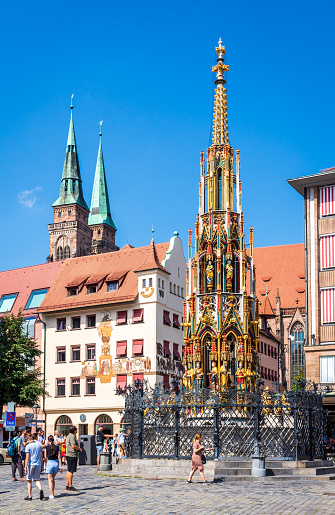 Nuremberg, Germany - August 23, 2023: General view of the 14th-century fountain Schöner Brunnen with Gothic spire on the main market, with the towers of St. Sebald church in the background.