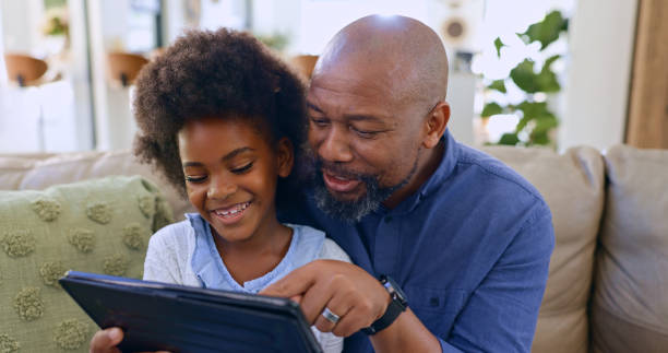 istock Dad, daughter and tablet with smile on sofa for games, streaming or elearning in living room of home. Black family, man and girl child with touchscreen, happiness or technology for watching movie 1987146720