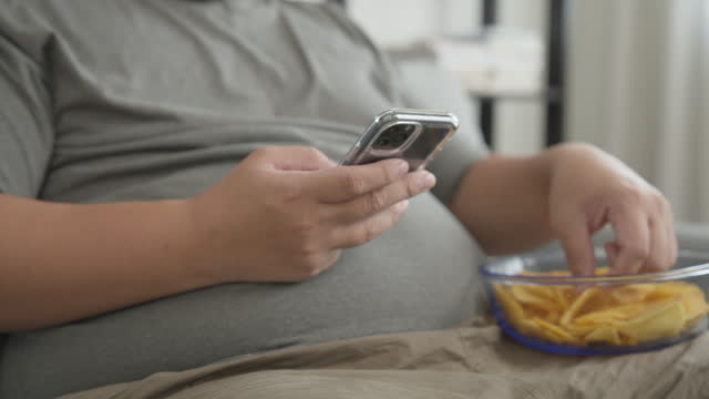 A big man enjoys eating potato chip snack while using a mobile phone