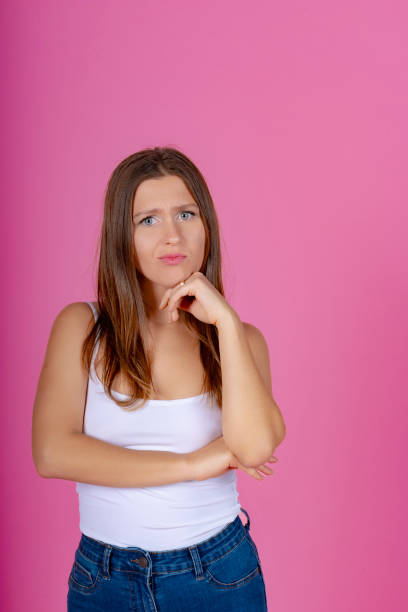 Thoughtful young woman in a white tank top with crossed arms, against a vivid pink background.