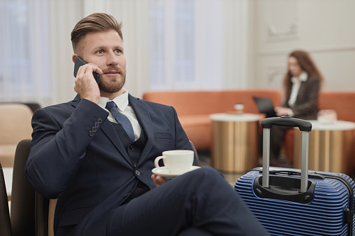 Portrait of elegant young businessman with suitcase speaking on phone in hotel lobby and enjoying cup of coffee, copy space