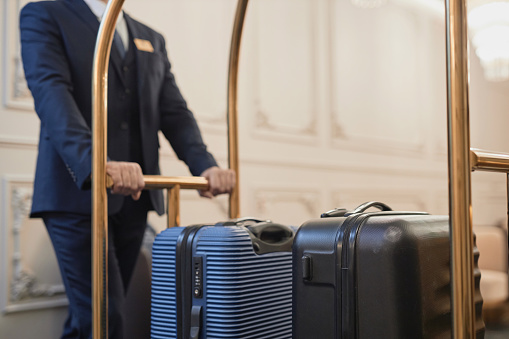Close up of male porter wearing suit pushing cart with bags and luggage in luxury hotel, copy space