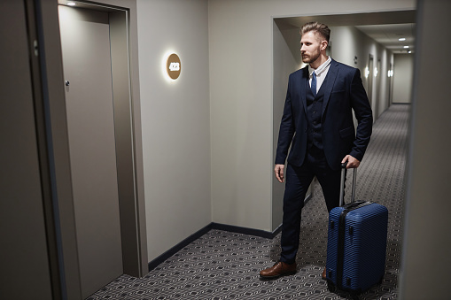 Full length portrait of handsome young businessman with suitcase walking to hotel room door in corridor, copy space