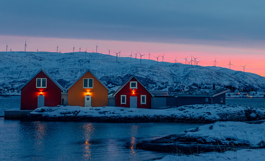 Small Houses standing near a small lake with the light reflecting in the water and wind turbines standing in the background and the sun setting in a beautiful orange color